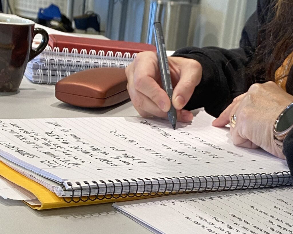 Närbild av en hand som skriver i ett kollegieblock. Runt omkring ligger stenciler, glasögonfodral och även en kaffekopp.