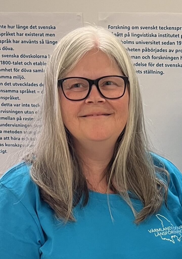 Victoria Jern Nilsson, Lokalvårdare Anna har långt silverfärgat hår och glasögon med tunna bågar. På bilden har hon på sig en turkos t-shirt med logotypen för Värmlands dövas länsförening