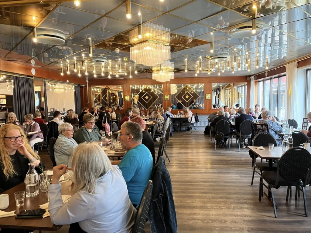 Människor sitter och äter vid flera småbord. I taket hänger guldfärgade lampor