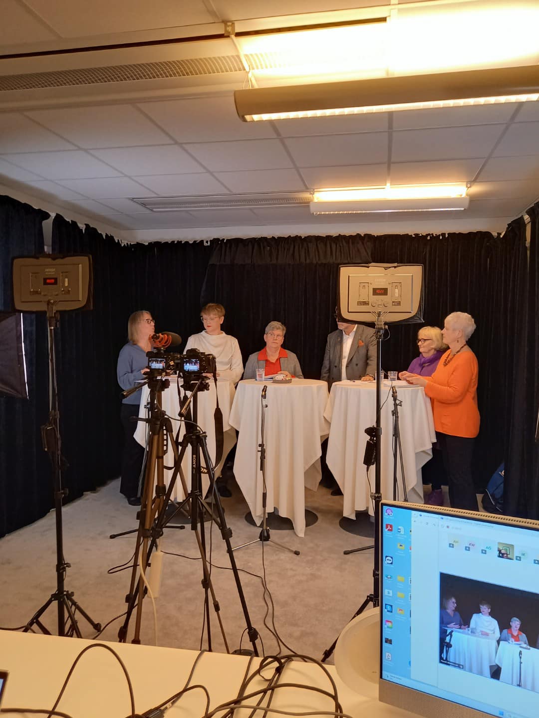 Sex personer runt ett avlångt bord. I förgrunden finns mikrofonstativ och en skärm där människorna syns i bild.