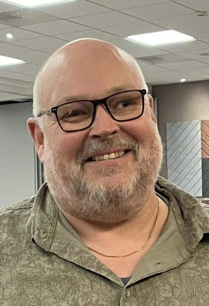 Stefan har rakat huvud, mustasch och helskägg. På bilden har han glasögon med svarta bågar och ett varmt leende.