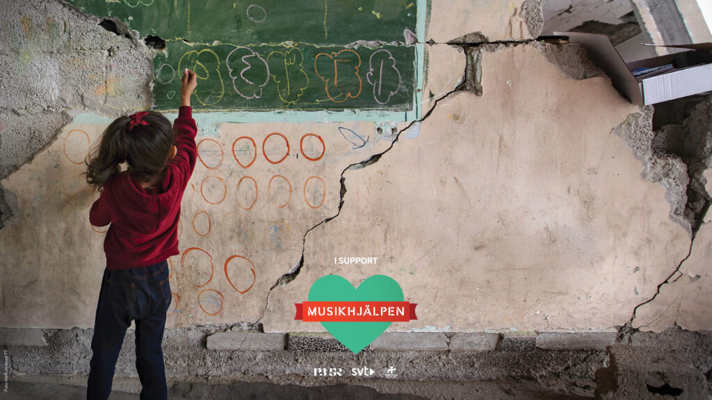 Ett mörkhårigt barn ritar med vit krita på en krön griffeltavla. Klassrummet utgörs av en betong-bunker