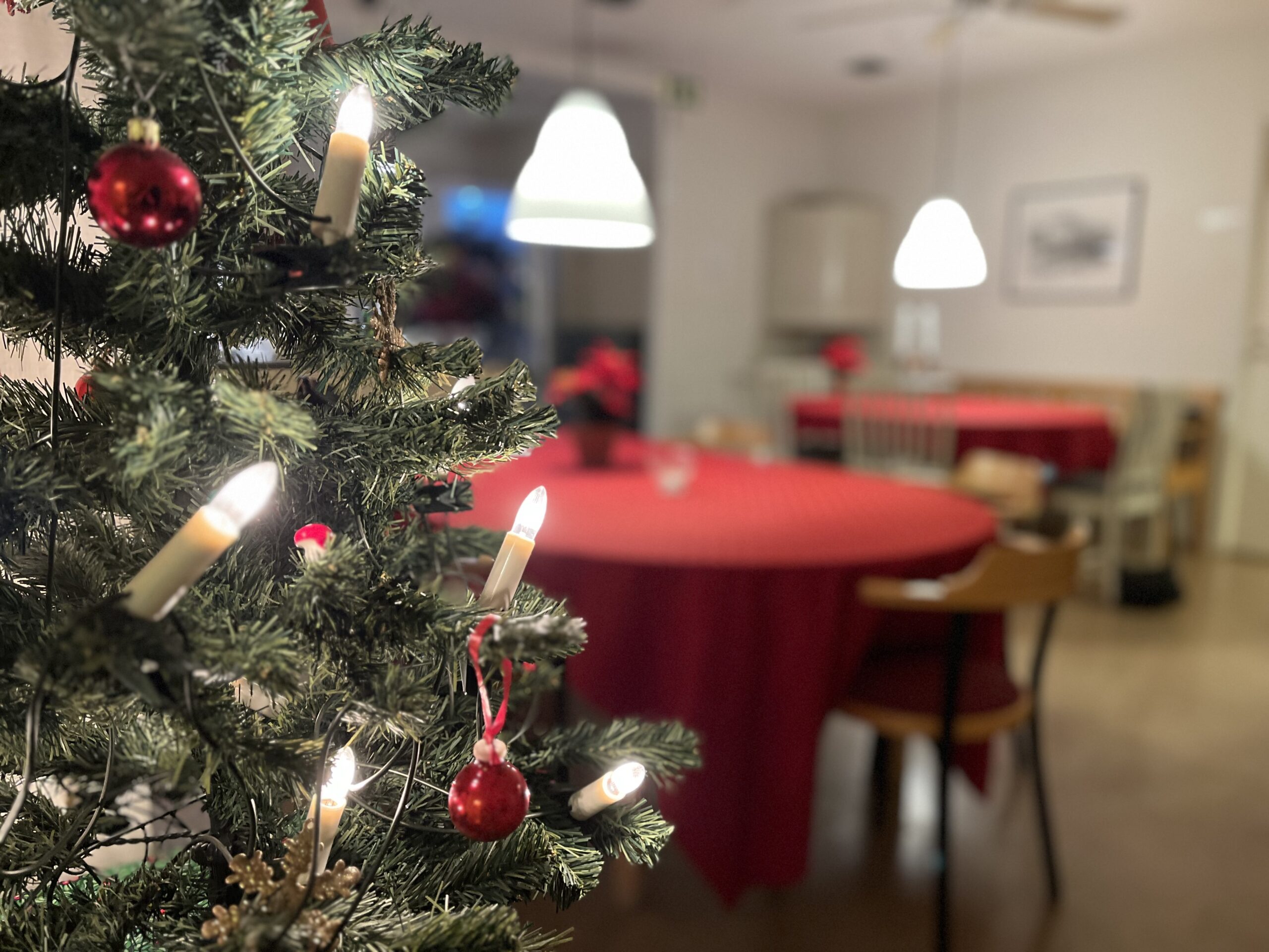 En liten plastgran med glänsande röda julkulor i förgrunden. I bakgrunden ett tomt fikarum med röda juldukar på borden.
