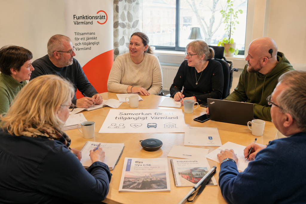 Personer med olika erfarenheter och funktionsnedsättningar sitter samlade runt ett mötesbord och diskuterar frågor som rör tillgänglighet och delaktighet i Värmland. På bordet ligger dokument om kollektivtrafik och Nya CSK. I bakgrunden syns Funktionsrätt Värmlands rollup.